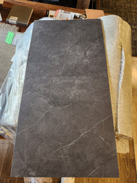 A Coastline Kamari (LVT) by Melmart Canada in dark gray with light veining sits beside a rug and flooring samples.