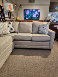 Superstyle 1014 Trinity Double Sofabed Sectional in beige with patterned pillows; TV shows pink tulips in background.