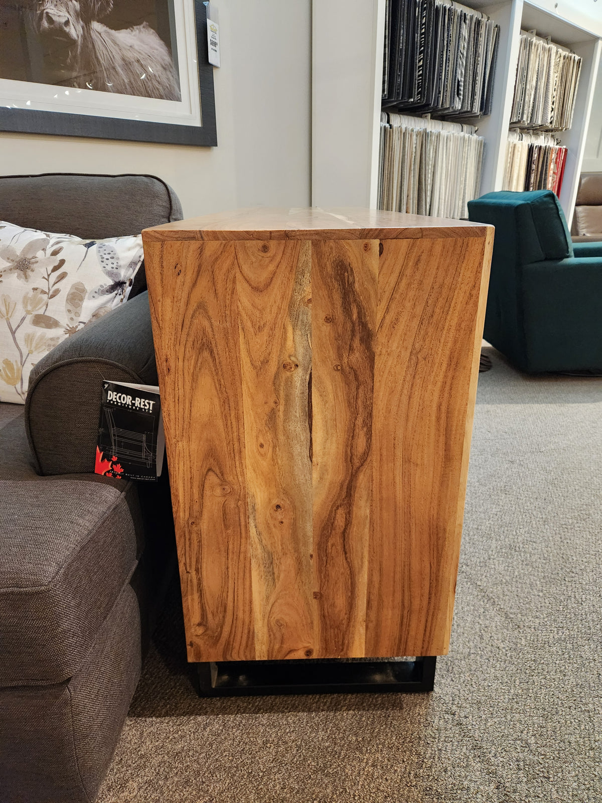The Jofran Geometrix Accent Cabinet with a natural grain finish stands by a gray sofa in a stylish showroom.