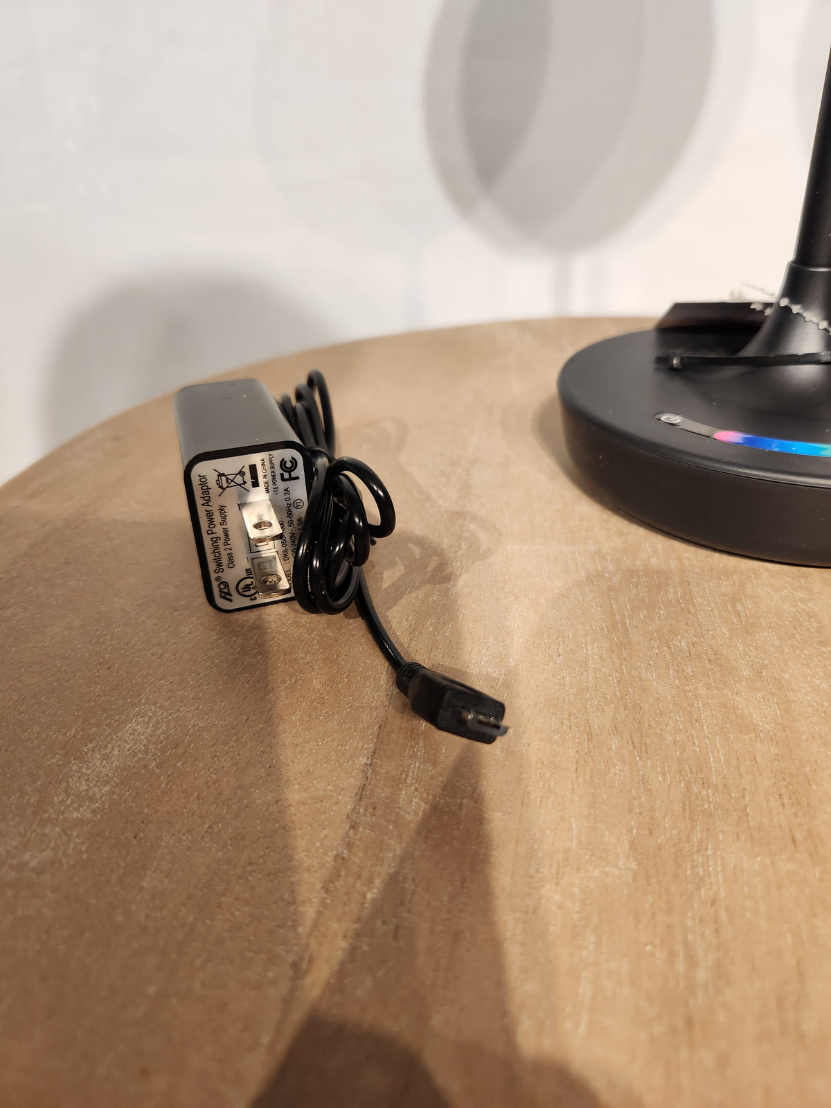 An Adesso Lamp Millie Color Changing Table Lamp sits on a wooden surface beside its black power adapter with cord.