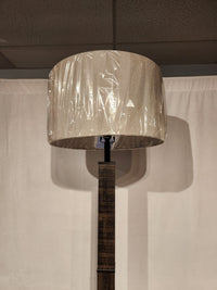 The Adesso Kona Floor Lamp features a beige shade and farmhouse style, standing tall in front of a white curtain.