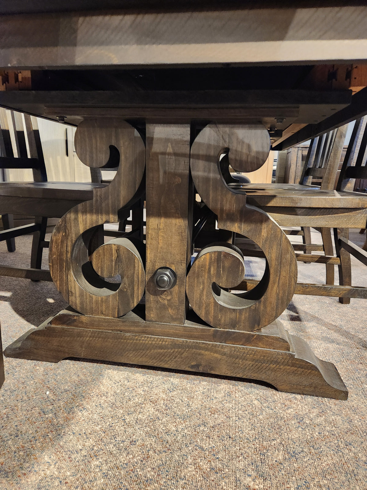 Close-up of the carved base on the 2491 Bellamy Dining Package by Magnussen, with chairs on a carpeted floor.