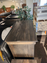 The Vokes 262-405 Bayshore 5 Drawer Chest is featured in a store, surrounded by mattresses and plants.