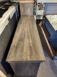 A wooden storage bench with a high backrest sits near a Vokes 262-407 Bayshore 7 Drawer Dresser in the showroom.