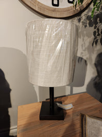 The Adesso Lamp Liam Table Lamp features a bronze finish and fabric shade, displayed on a wooden surface.