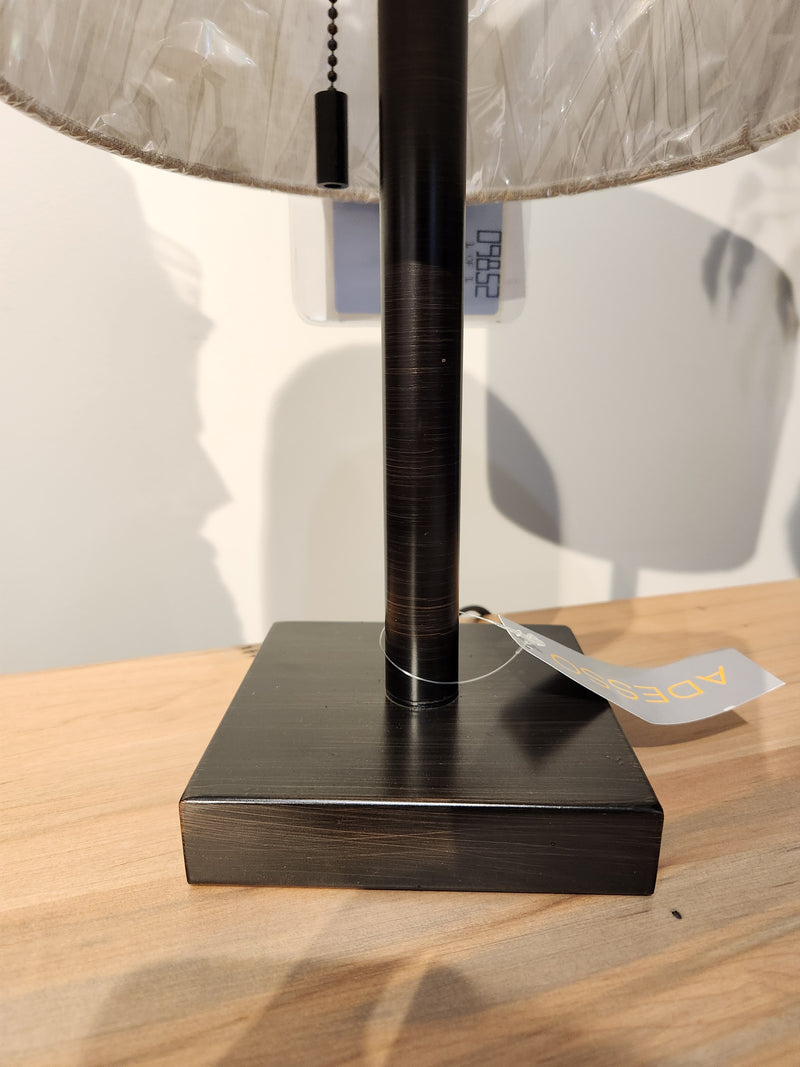 Close-up of Adesso Lamp’s Liam Table Lamp with bronze base, wood surface, price tag, and plastic-wrapped shade.