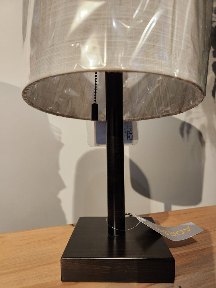 The Adesso Lamp Liam Table Lamp with a square black base and fabric shade sits on a wooden surface.
