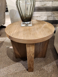 Magnussen T6348 Hayden Round End Table in Golden Oat finish with vase, on carpet near wood-paneled wall.