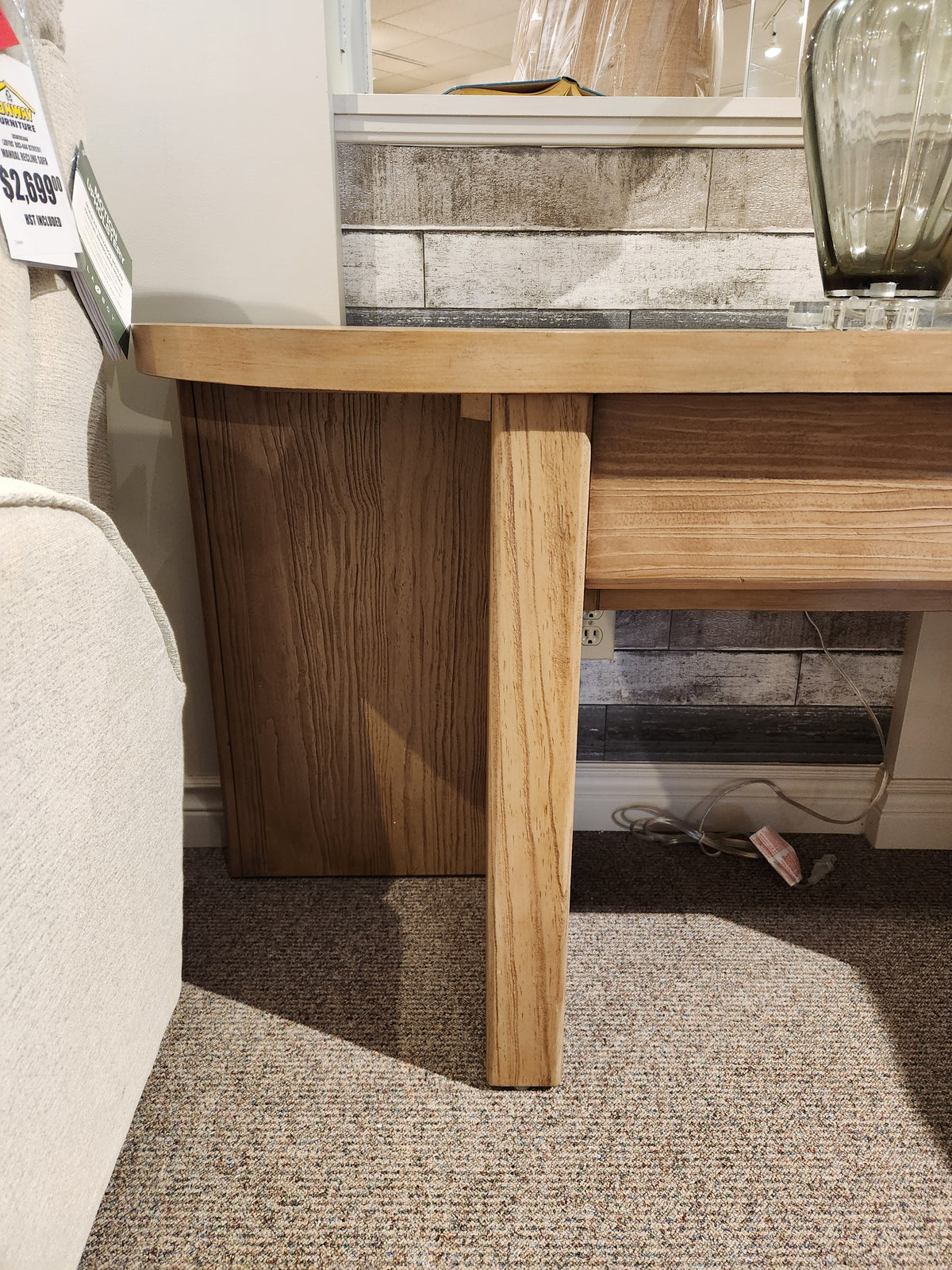 The Magnussen T6348 Hayden Sofa Table in Golden Oat stands by a beige couch on a carpeted floor with cables underneath.