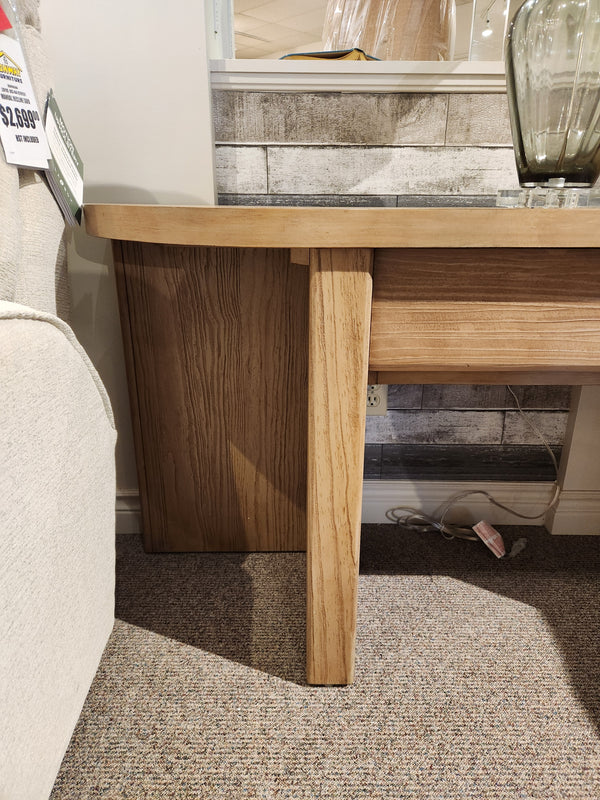 The Magnussen T6348 Hayden Sofa Table in Golden Oat stands by a beige couch on a carpeted floor with cables underneath.