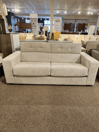 The Donald Choi Edison Queen Sofa Bed, beige with tufted back and hardwood frame, shown in a furniture showroom.