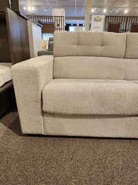 The Donald Choi Edison Queen Sofa Bed, beige with one armrest, is displayed on a brown carpet in the showroom.