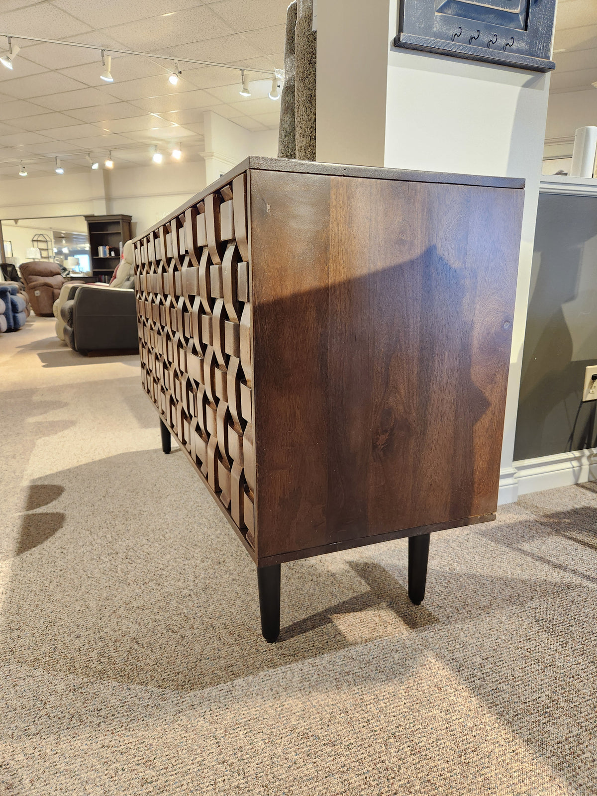 The Jofran Trent Woven Front Accent Cabinet 60" features a geometric design and black legs, shown on carpeted flooring.