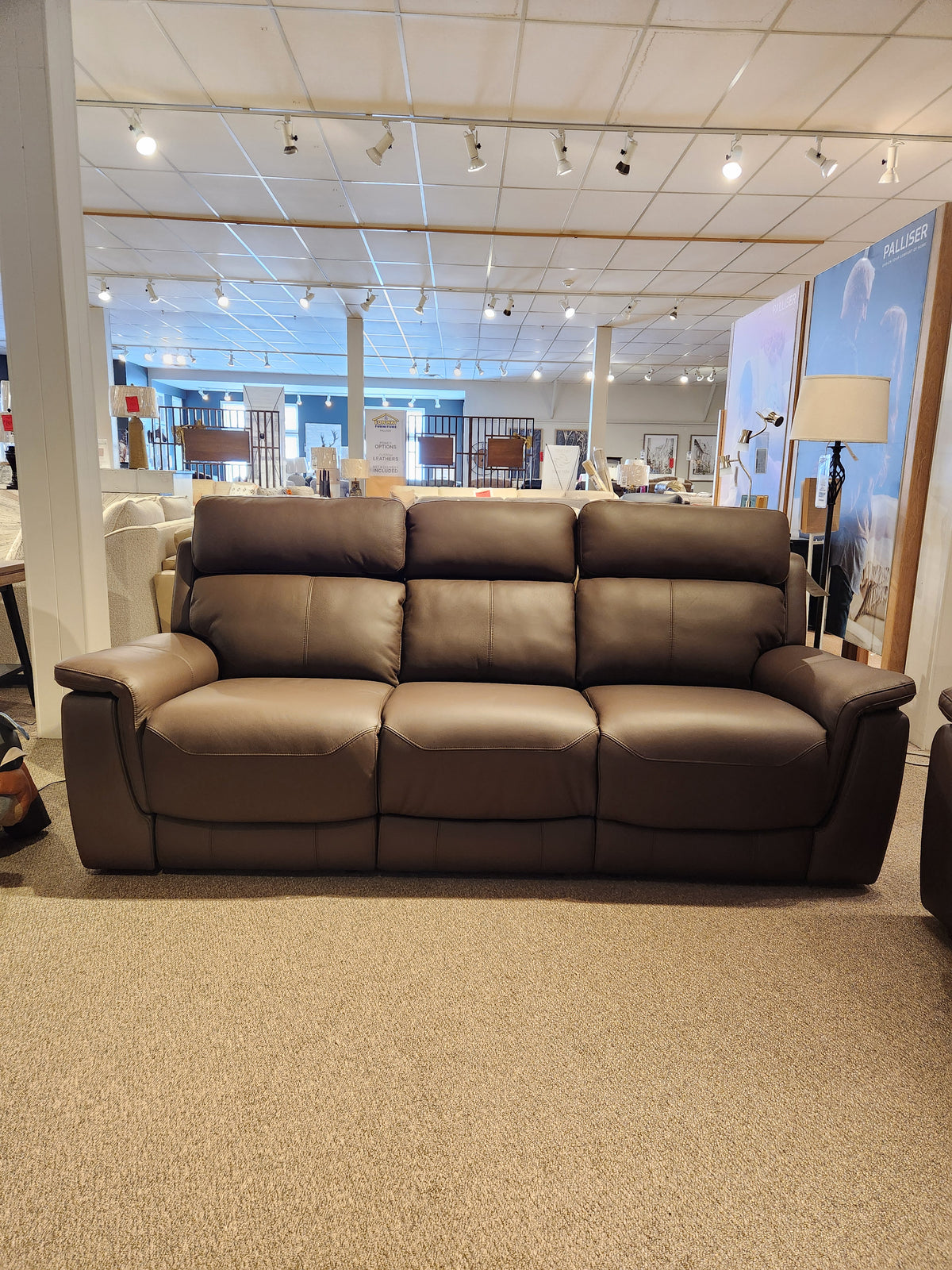 The Palliser 41051 Henry Sofa, a brown leather power recliner, is displayed in a carpeted showroom with overhead lights.