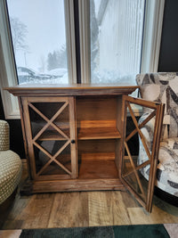 The Jofran 2028 Carrington Small Breakfront Cabinet with glass doors sits between chairs by a window on a snowy day.