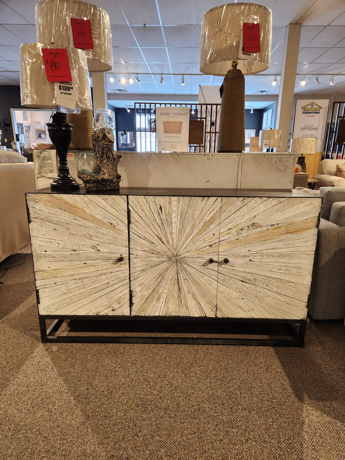 The Jofran 1929 Astral Plains 3 Door Cabinet features starburst doors and is shown in a showroom with lamps on top.