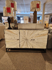 The Jofran 1929 Astral Plains 3 Door Cabinet features starburst doors and is shown in a showroom with lamps on top.