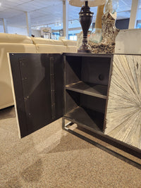 The Jofran 1929 Astral Plains 3 Door Cabinet has textured doors, black metal reveals, and two shelves on a carpeted floor.