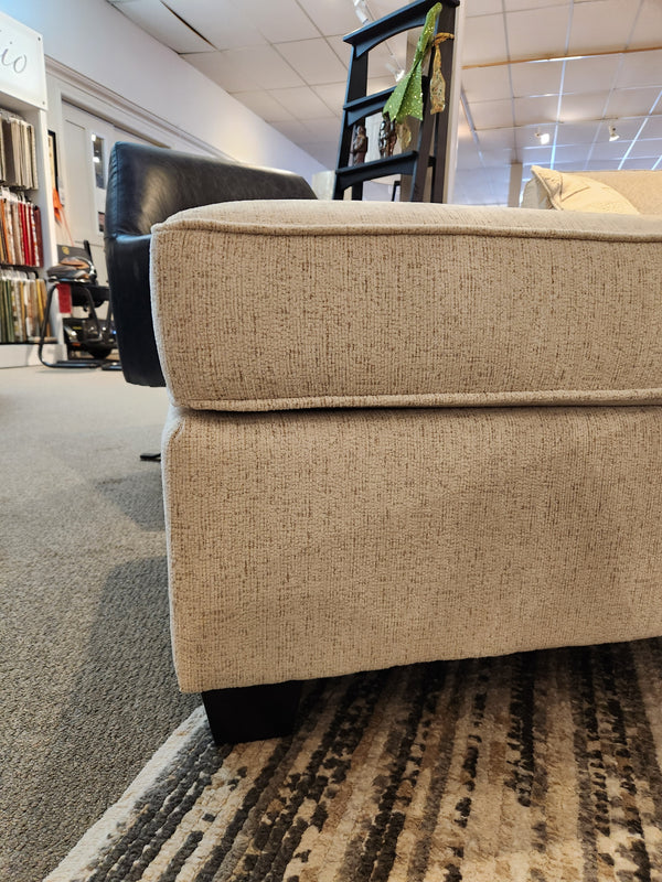 A close-up side view of a Decor-rest 2541 Ottoman next to a beige sofa on a patterned rug in a showroom.