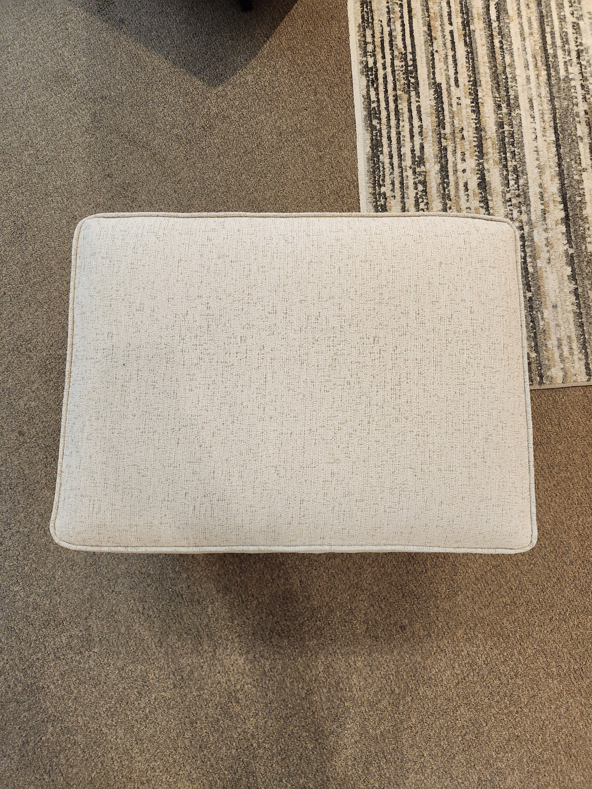 The Decor-rest 2541 Ottoman acts as a footrest or seat on a brown carpet beside a striped area rug, shown from above.