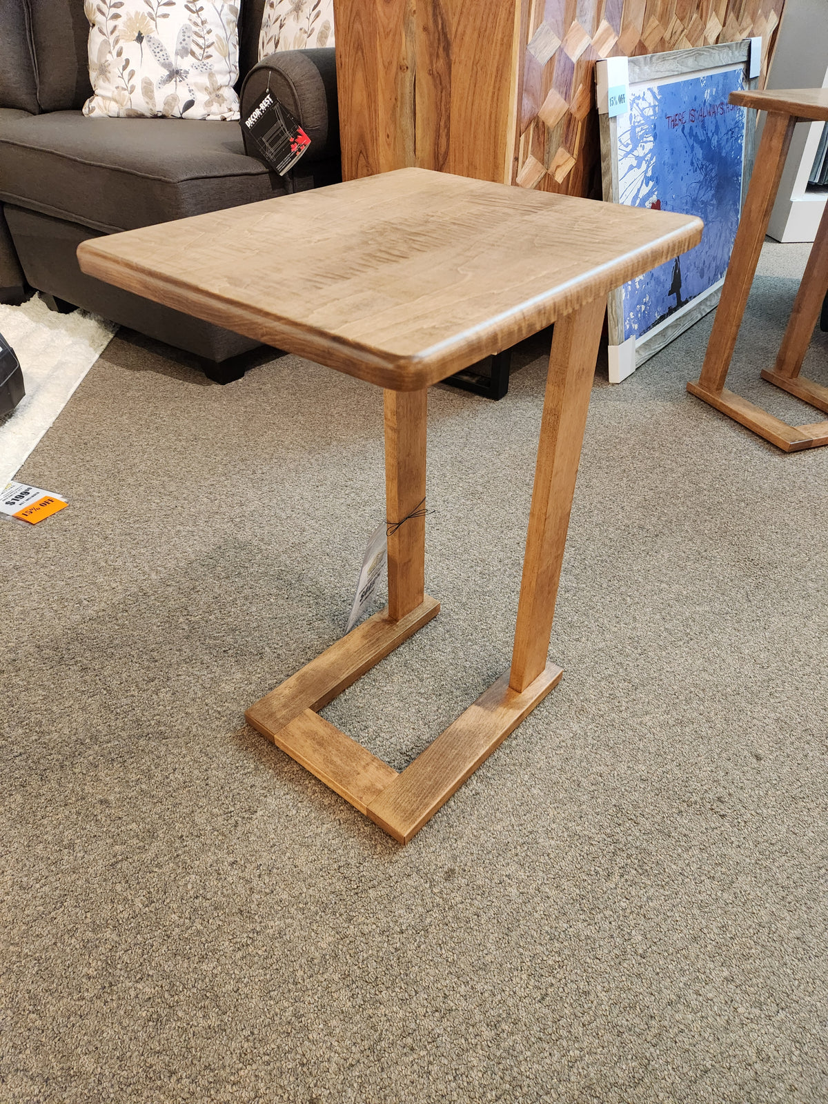 The Handstone AlaCarte Outback Snack Table, a small wooden C-shape, sits on carpet in a furniture store.