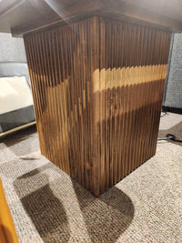 The Handstone Lotus End Table with elegant grooves stands near a gray couch and wall on a carpeted floor.