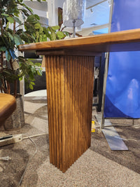 The Lotus Sofa Table by Handstone features vertical grooved legs, shown on carpet near a plant and blue divider.