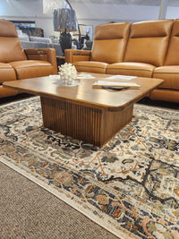 The Handstone Lotus Coffee Table sits on a patterned rug, surrounded by tan leather recliners in a showroom.