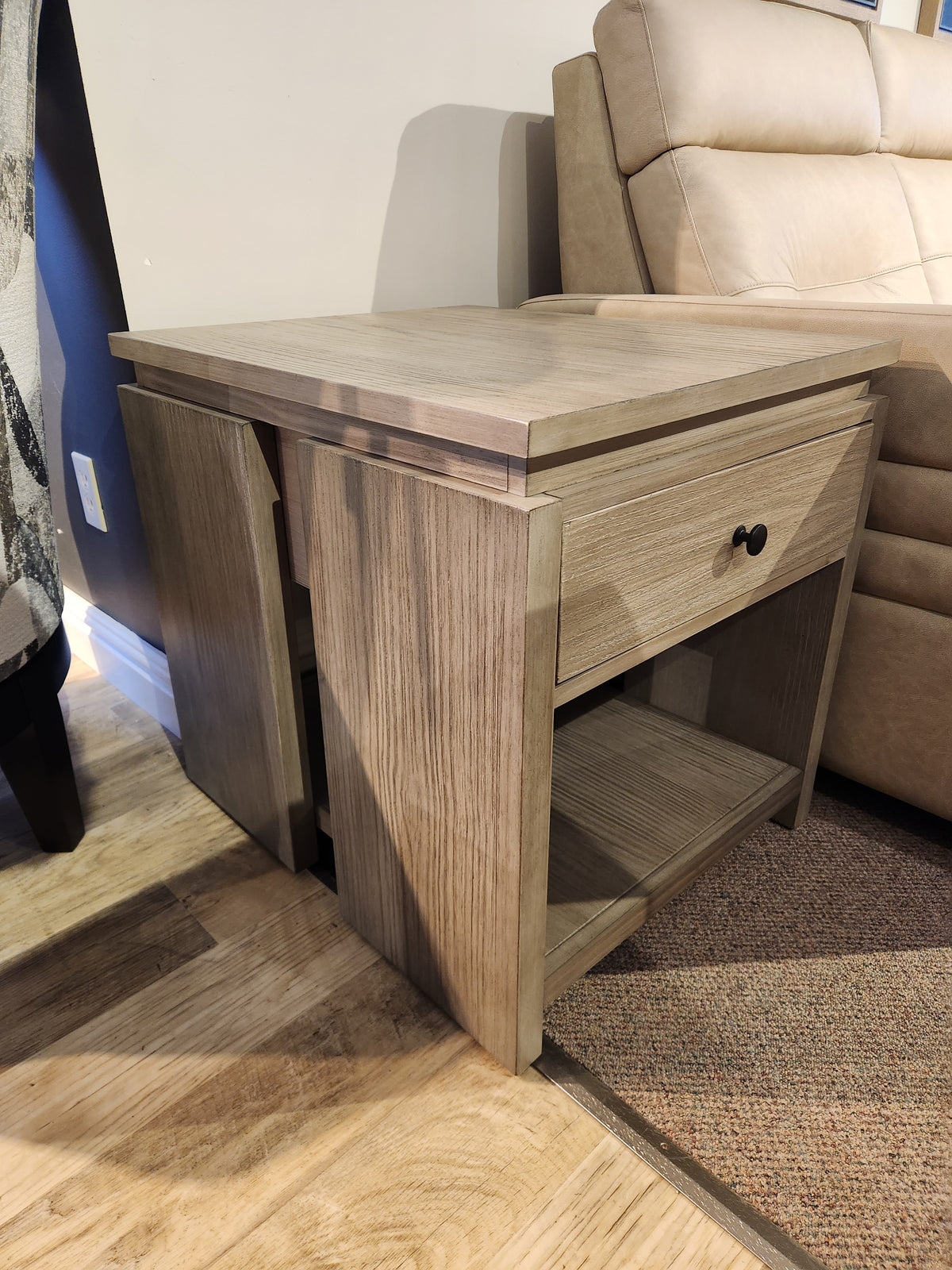 The T5865 Conrad End Table by Magnussen in Stone Harbor Grey with bronze hardware sits beside a beige sofa.