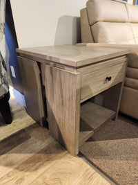 The T5865 Conrad End Table by Magnussen in Stone Harbor Grey with bronze hardware sits beside a beige sofa.