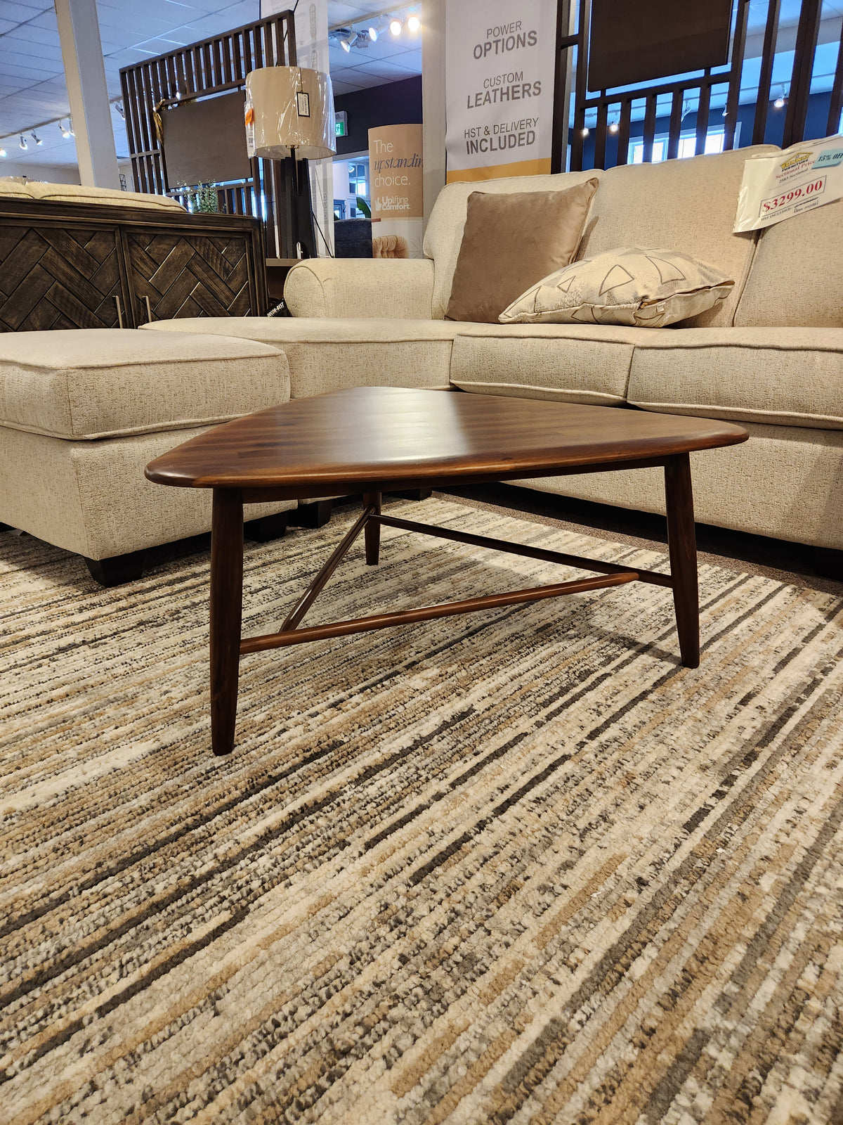 The 7110 Kacia Tri Coffee Table by Palliser sits on a striped rug before a beige sectional sofa in the showroom.