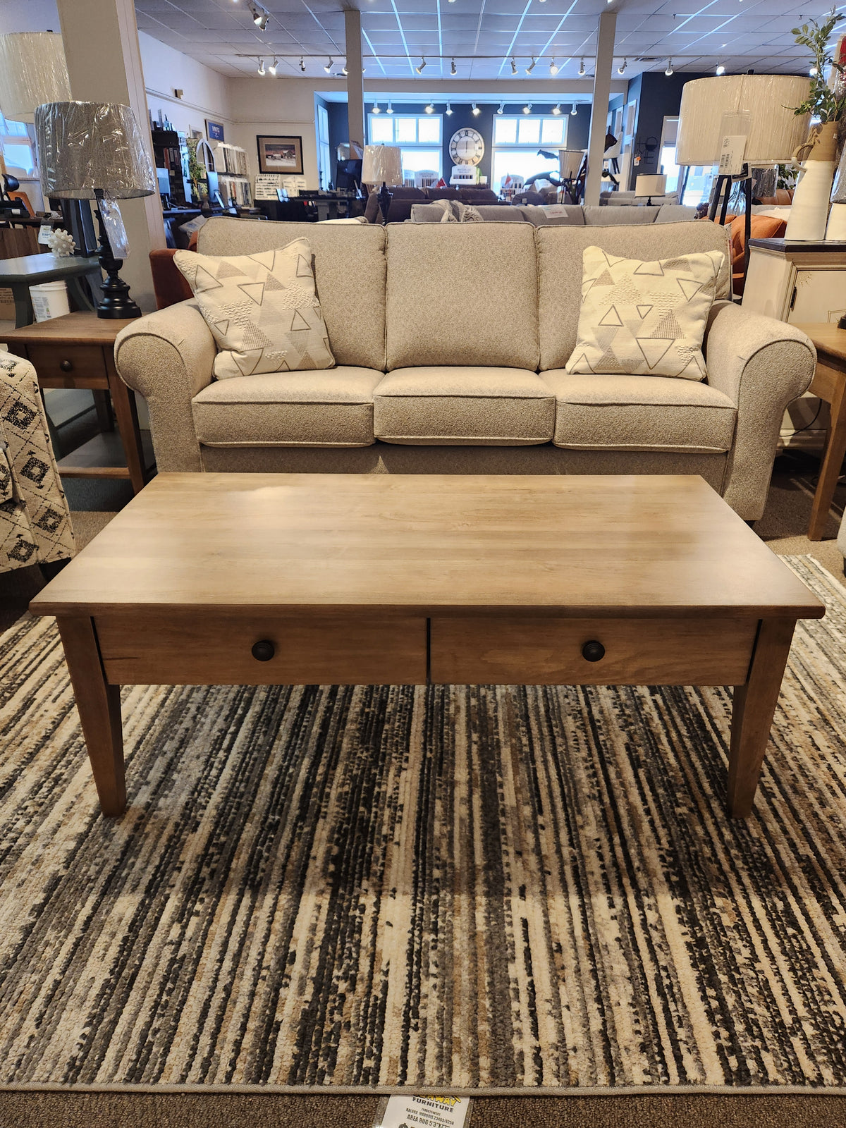 A beige sofa with patterned pillows sits behind a Durham 905-2 Drawer Champagne Cocktail Table on a striped rug.