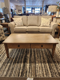 A beige sofa with patterned pillows sits behind a Durham 905-2 Drawer Champagne Cocktail Table on a striped rug.