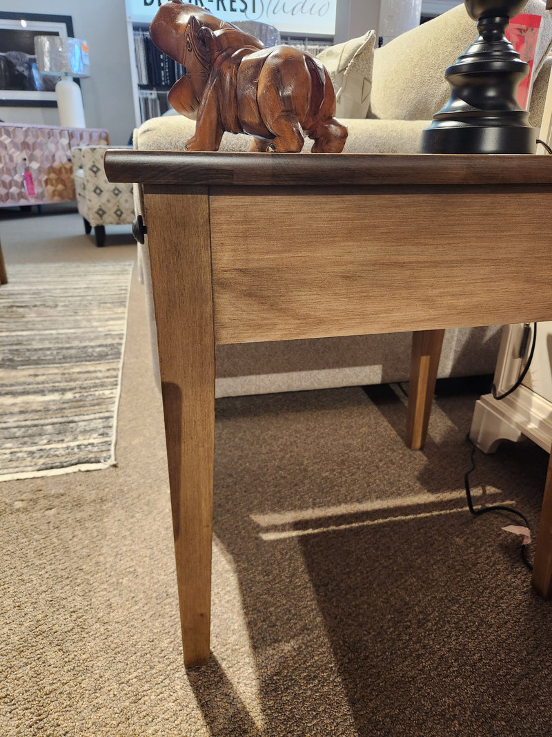 A wooden elephant figurine sits on a Durham 905 20"x24" Champagne end table with drawer beside a beige sofa and black lamp.