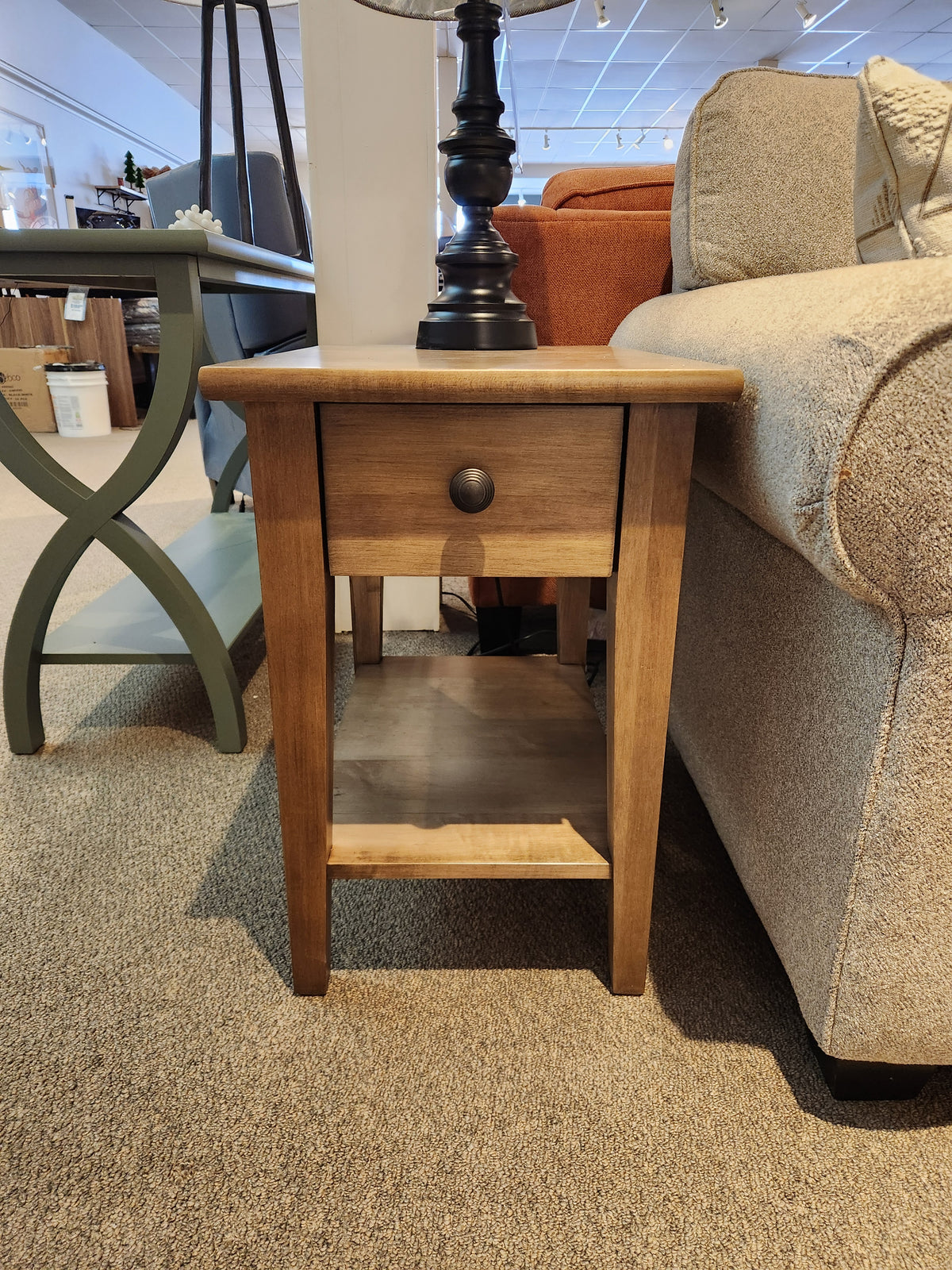 A Durham 905 16"x24" Champagne end table with a drawer sits between a beige sofa and green side table in store.