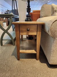 A Durham 905 16"x24" Champagne end table with a drawer sits between a beige sofa and green side table in store.