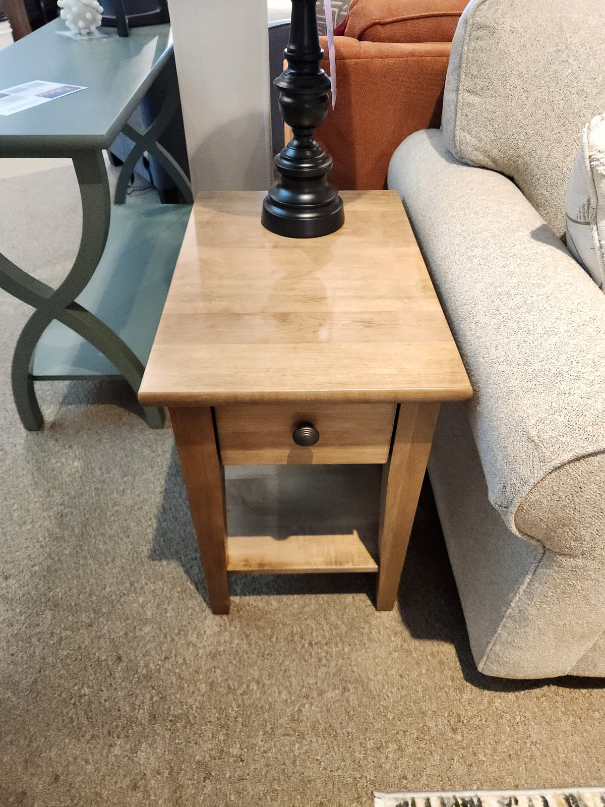 A Durham 16"x24" Champagne end table with drawer sits between a beige sofa and green accent table.