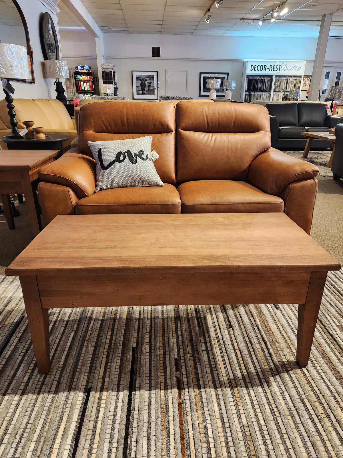 Brown leather loveseat with a "Love" pillow behind Durham's 905 Cocktail Table (48x24", Aged Wheat) on a striped rug.