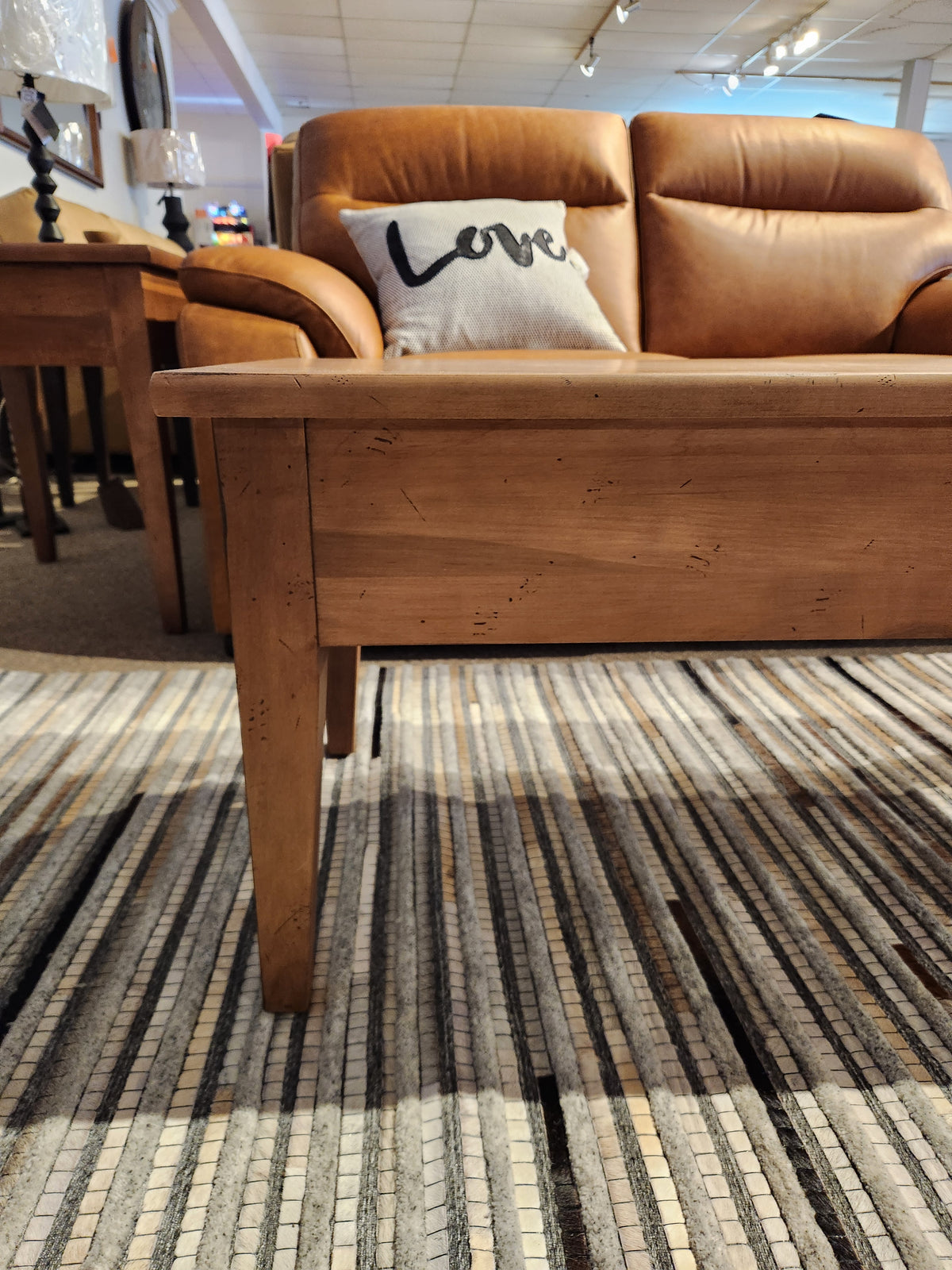 Durham 905-48 x 24" Cocktail Table in Aged Wheat displayed before a brown leather sofa with "Love" pillow on striped rug.
