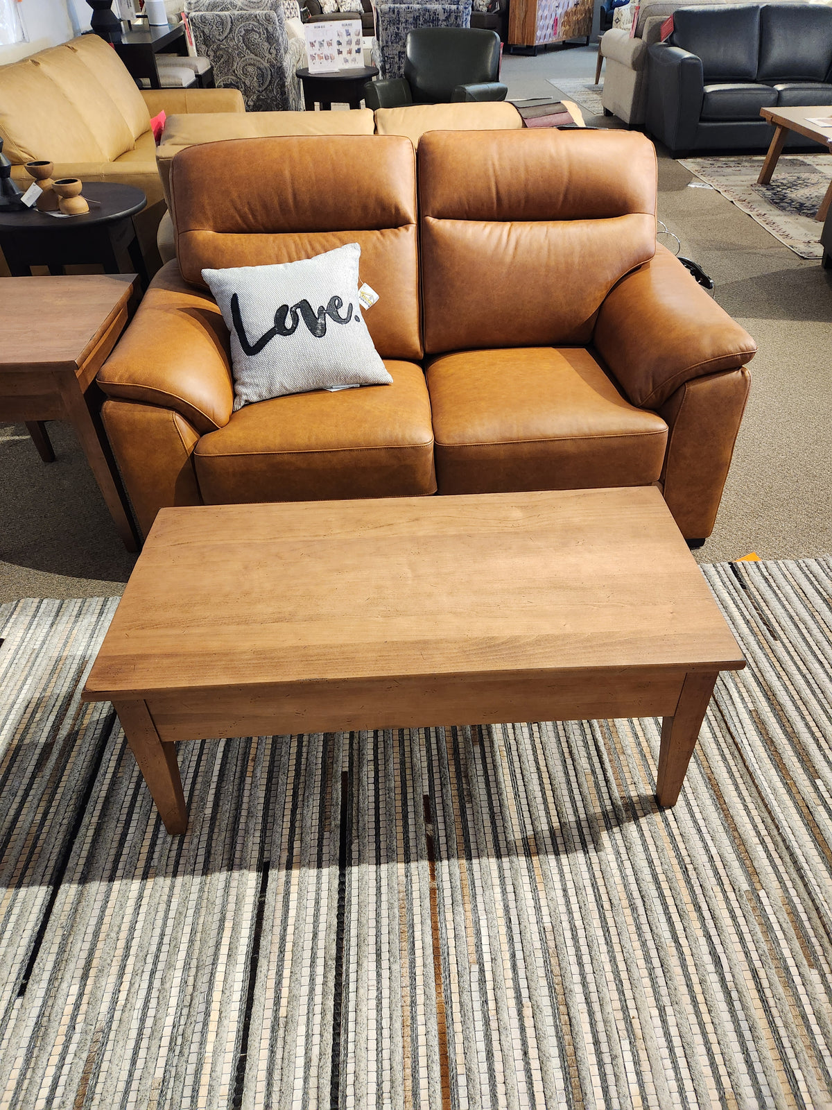 A brown leather loveseat with a "Love." pillow sits behind a Durham 905 Aged Wheat cocktail table on a striped rug.