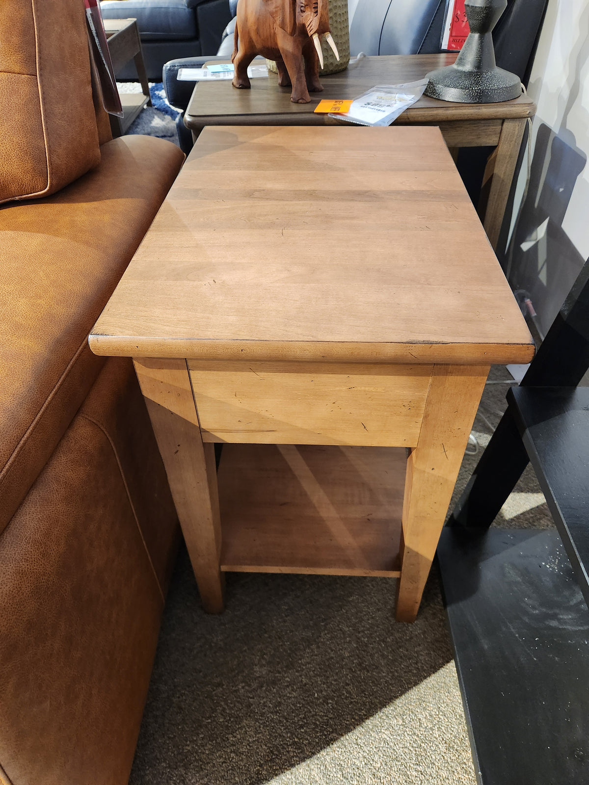 The Durham 905 16" x 24" End Table in Aged Wheat is displayed between a brown couch and black shelving in-store.