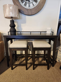 The Durham 905 Console Table with metal rail includes two stools, topped with a black lamp and white speaker under a wall clock.