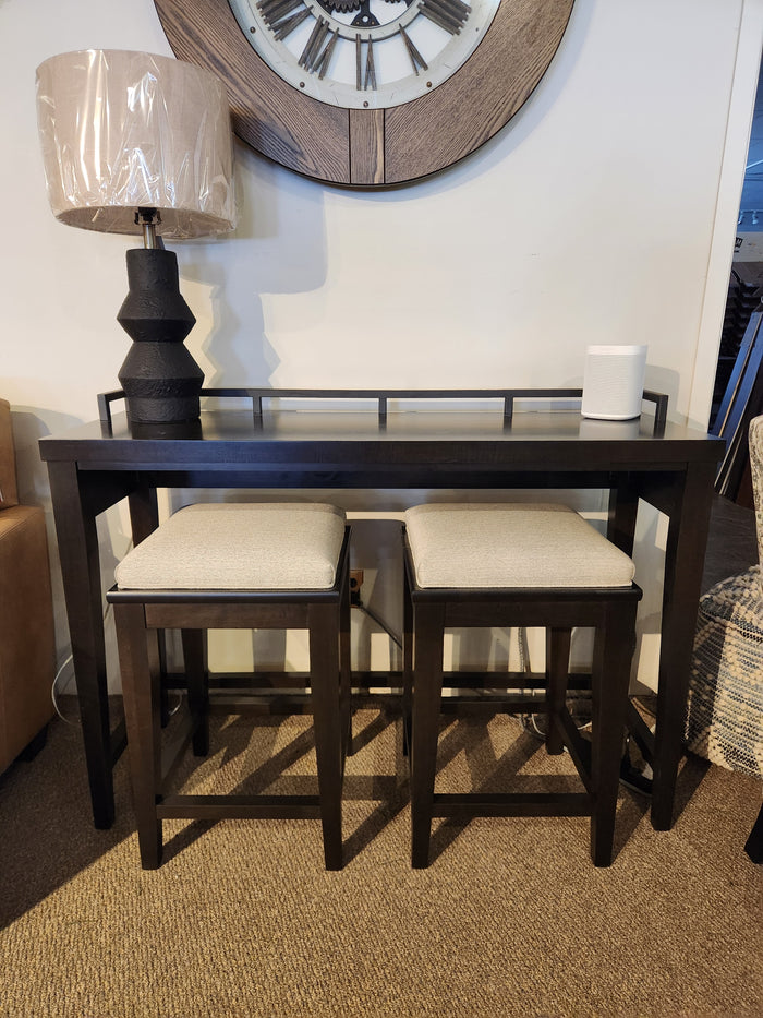 The Durham 905 Console Table with metal rail includes two stools, topped with a black lamp and white speaker under a wall clock.