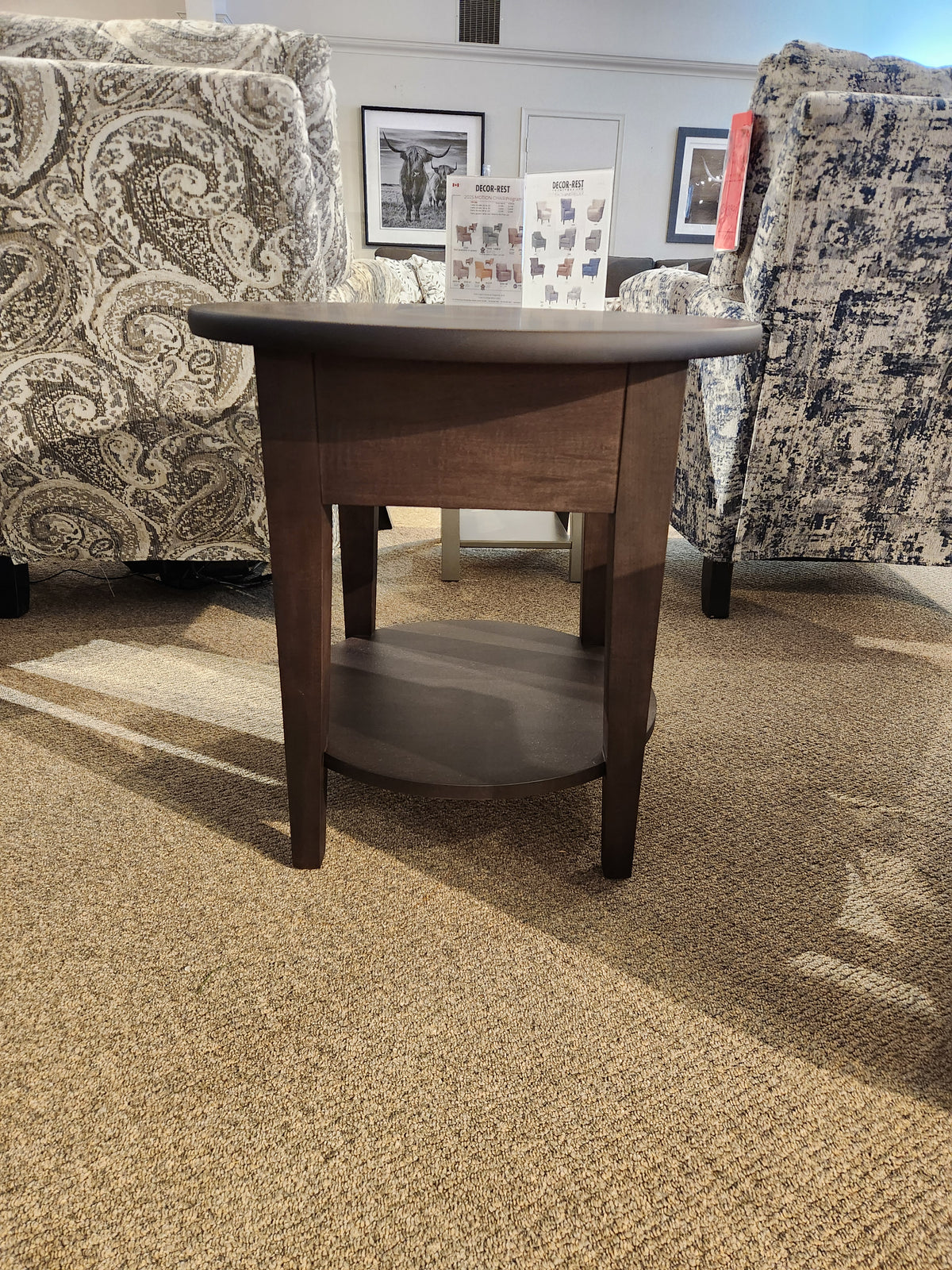 The Durham 905-24" Round Table w/Shelf in Java rests between patterned armchairs on a carpeted floor.