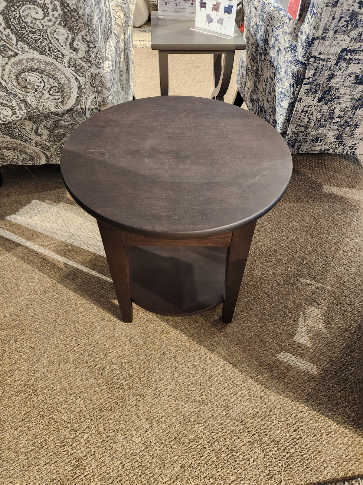 The Durham 905-24" Round Table with shelf in Java provides storage and is shown on a beige carpet in the showroom.