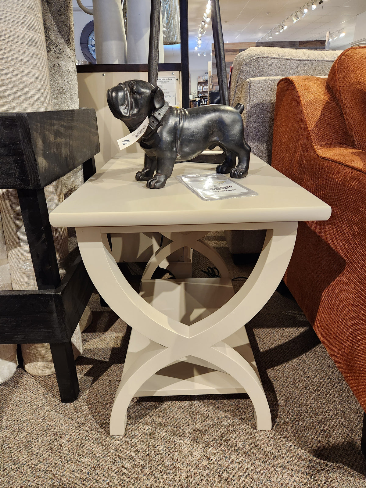 The Durham 900-534 Transitional End Table features a beige finish and curved X-base, shown with a black dog figurine.