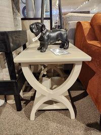 The Durham 900-534 Transitional End Table features a beige finish and curved X-base, shown with a black dog figurine.