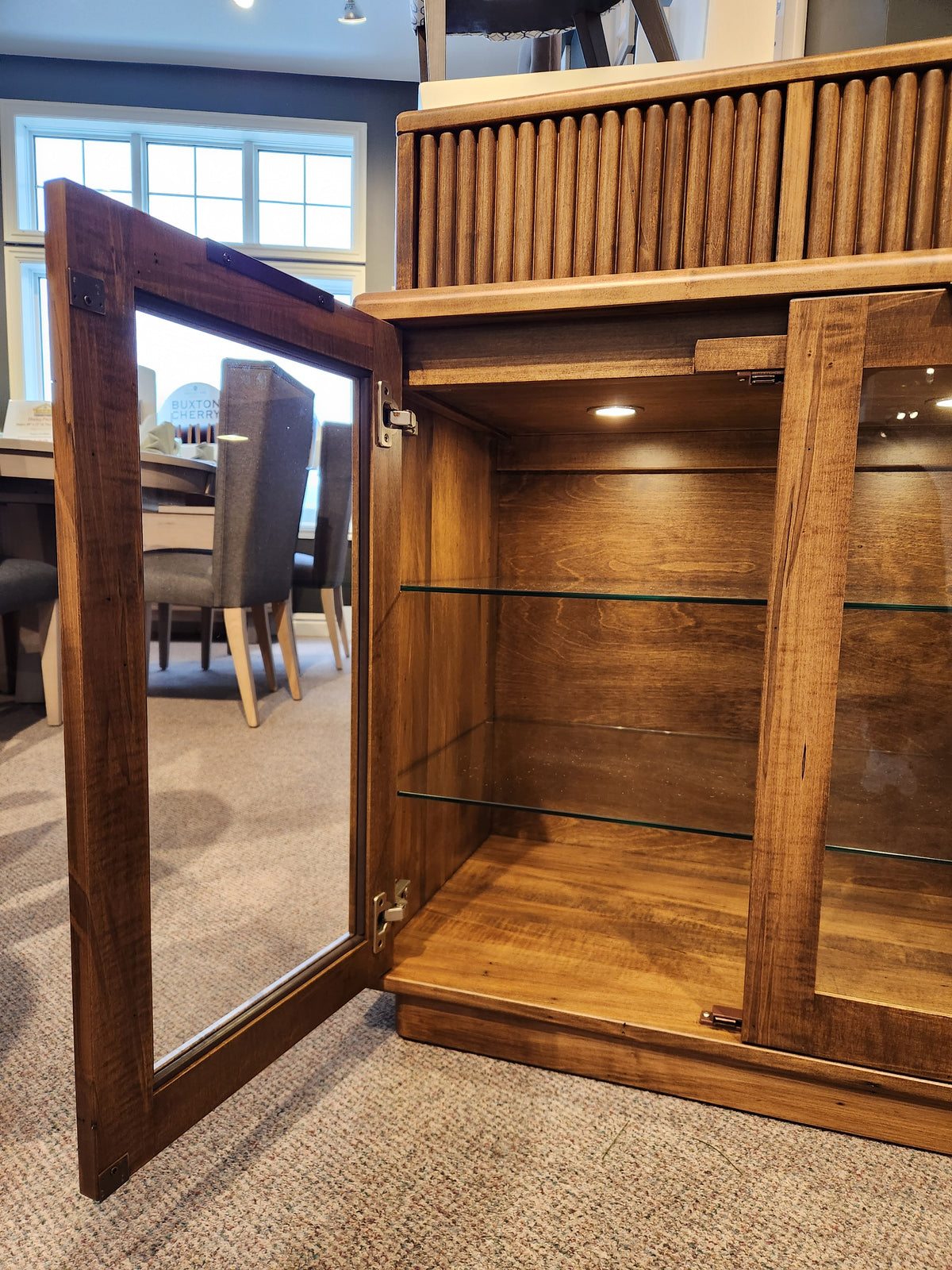 Glass door cabinet with wooden shelves open, shown in a room with carpet and Handstone Lotus Dining Sideboard with pot lights.