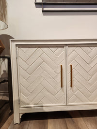 Jofran Gramercy 3 Door Accent Cabinet with chevron inlay and gold handles, shown on a wooden floor.