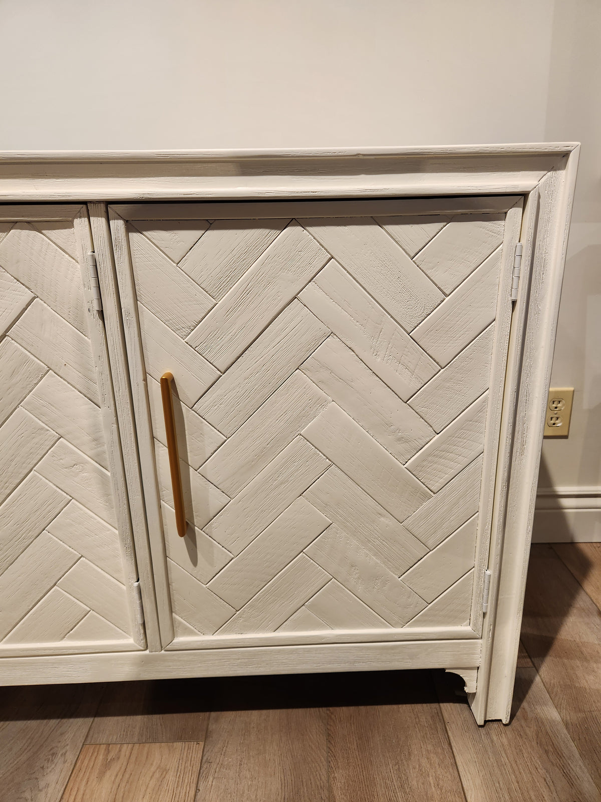 The Jofran Gramercy 3 Door Accent Cabinet features a chevron inlay, gold handles, and sits on a light wood floor.